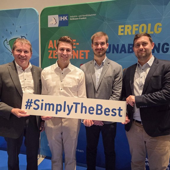 Bestenehrung Tim Leiser Vor einem Messehintergrund stehen 4 Männer. Alle sind schick angezogen und tragen Hemd oder Jackett. Sie lächeln in die Kamera und halten ein Schild mit #simplythebest in die Kamera.