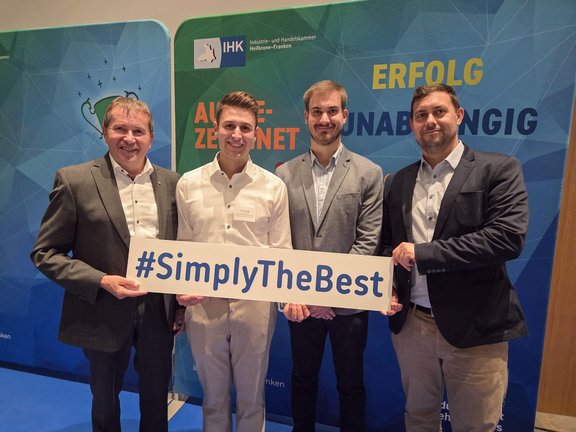 Bestenehrung Tim Leiser Vor einem Messehintergrund stehen 4 Männer. Alle sind schick angezogen und tragen Hemd oder Jackett. Sie lächeln in die Kamera und halten ein Schild mit #simplythebest in die Kamera.