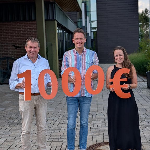 Drei Personen vor einem modernen Gebäude präsentieren einen symbolischen Spendenscheck in Höhe von 10.000 Euro, dargestellt durch große orangefarbene Zahlen und ein Eurozeichen.