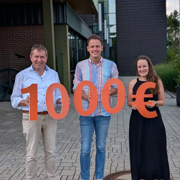 Drei Personen vor einem modernen Gebäude präsentieren einen symbolischen Spendenscheck in Höhe von 10.000 Euro, dargestellt durch große orangefarbene Zahlen und ein Eurozeichen.