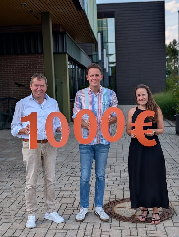 Spendenübergabe Lebenshilfe Drei Personen vor einem modernen Gebäude präsentieren einen symbolischen Spendenscheck in Höhe von 10.000 Euro, dargestellt durch große orangefarbene Zahlen und ein Eurozeichen.