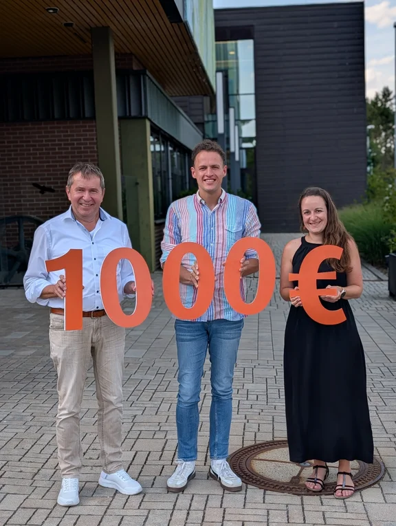 Drei Personen vor einem modernen Gebäude präsentieren einen symbolischen Spendenscheck in Höhe von 10.000 Euro, dargestellt durch große orangefarbene Zahlen und ein Eurozeichen.