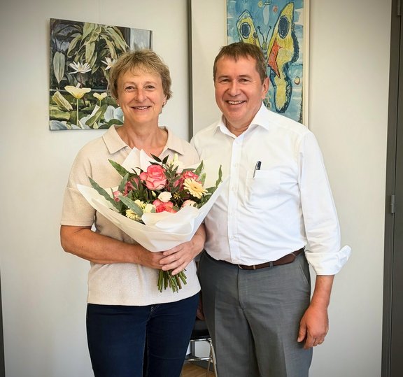 Eine Person hält einen Blumenstrauß mit rosa und weißen Blumen in den Händen und steht gemeinsam mit einer weiteren Person in einem Büro. Beide blicken in die Kamera. Im Hintergrund sind zwei gerahmte Kunstwerke an einer hellen Wand zu sehen. Die Aufnahme entstand anlässlich eines Mitarbeiterjubiläums.