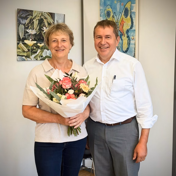 Eine Person hält einen Blumenstrauß mit rosa und weißen Blumen in den Händen und steht gemeinsam mit einer weiteren Person in einem Büro. Beide blicken in die Kamera. Im Hintergrund sind zwei gerahmte Kunstwerke an einer hellen Wand zu sehen. Die Aufnahme entstand anlässlich eines Mitarbeiterjubiläums.