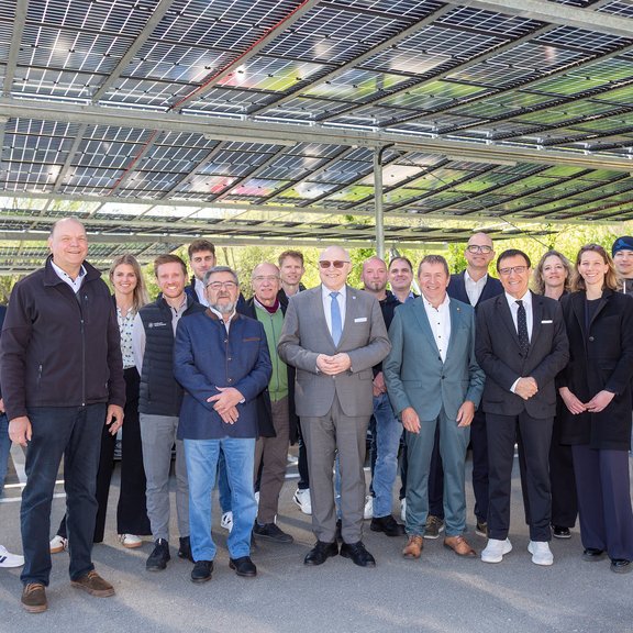Gruppe von Mitarbeitenden und Projektbeteiligten steht unter einer überdachten Parkplatzanlage mit installierten Photovoltaikmodulen