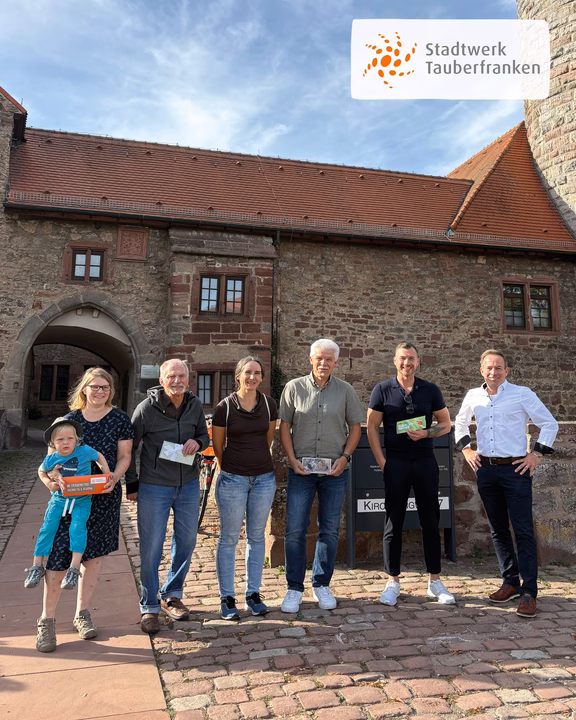 Gewinner Großer Markt 2026 Im Mittelpunkt steht eine Gruppe von sechs Erwachsenen und einem Kind vor einem historischen Steingebäude. Mehrere Personen halten Karten bzw. Gutscheine; oben rechts ist das Logo „Stadtwerk Tauberfranken“ eingeblendet.