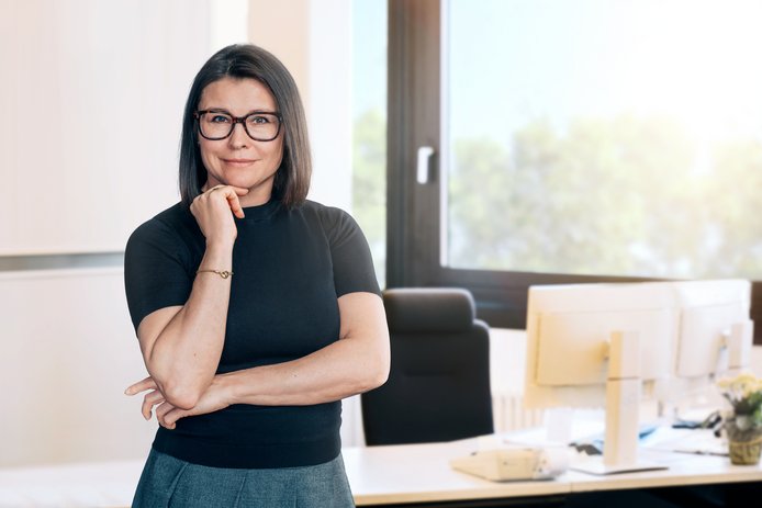 Eine Frau mittleren Alters steht vor einem Schreibtisch in einem Büro und lächelt in die Kamera. Sie trägt ein schwarzes Oberteil, hat schulterlange dunkle Haare und trägt eine Brille mit dunklen Rändern. 