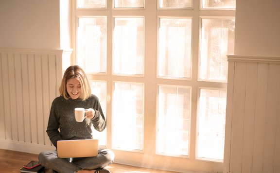 Zählerstände Eine junge Frau sitzt entspannt im Schneidersitz auf dem Boden vor einem großen Fenster, durch das warmes Sonnenlicht strömt. Sie hält eine Tasse in der Hand und blickt lächelnd auf ihren Laptop. Neben ihr liegen einige Notizbücher und ein Smartphone.