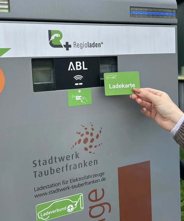 Eine Hand hält eine Ladekarte an ein Kartenlesegerät an einer E-Ladesäule mit der Aufschrift „ABL“ und dem Logo des Stadtwerks Tauberfranken.