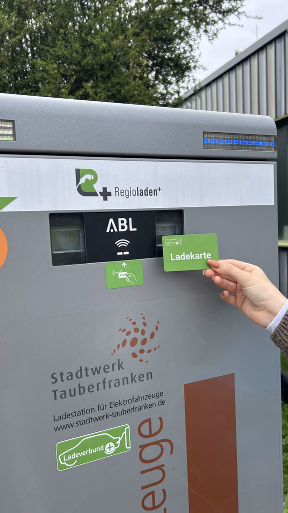 Eine Hand hält eine Ladekarte an ein Kartenlesegerät an einer E-Ladesäule mit der Aufschrift „ABL“ und dem Logo des Stadtwerks Tauberfranken.