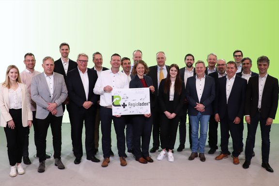 Eine Gruppe von etwa 20 Personen in formeller Kleidung steht vor einem hellgrünen Hintergrund. Zwei Personen in der Mitte halten ein Schild mit der Aufschrift 'Regioladen+' und mehreren Unterschriften, das das Datum der Gründung, den 16.01.2025, zeigt.