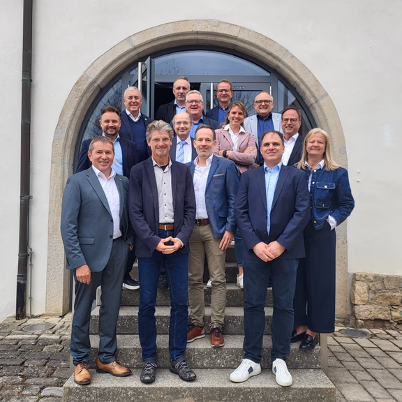 Mehrere Personen stehen in Gruppenanordnung auf einer Steintreppe vor einem Gebäude mit Rundbogen-Eingang und Glastür, aufgenommen bei einem Treffen des Regionalbeirats.