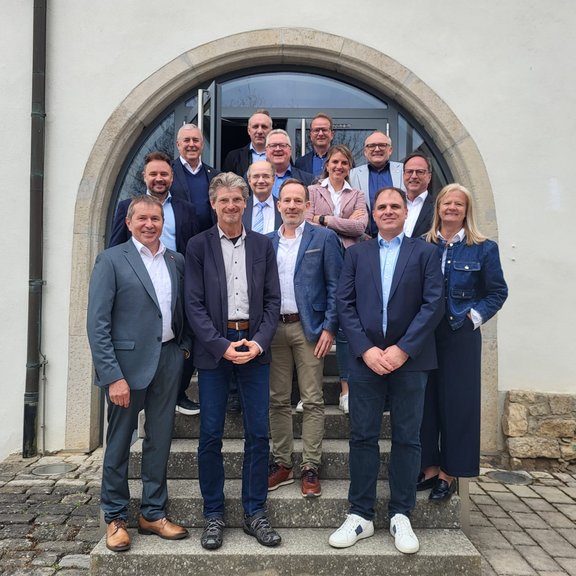 Mehrere Personen stehen in Gruppenanordnung auf einer Steintreppe vor einem Gebäude mit Rundbogen-Eingang und Glastür, aufgenommen bei einem Treffen des Regionalbeirats.