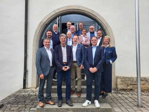 Mehrere Personen stehen in Gruppenanordnung auf einer Steintreppe vor einem Gebäude mit Rundbogen-Eingang und Glastür, aufgenommen bei einem Treffen des Regionalbeirats.