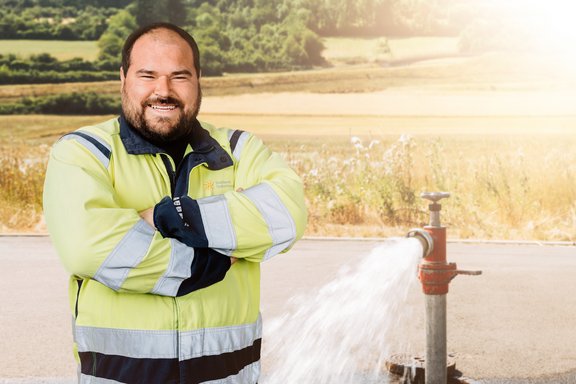 Ein Mann steht in gelber Arbeitskleidung links im Bild. Rechts ist ein Standrohr aufgebaut, aus dem Wasser sprudelt. Der Hintergrund sind sommerliche Getreidefelder mit einzelnen Bäumen.