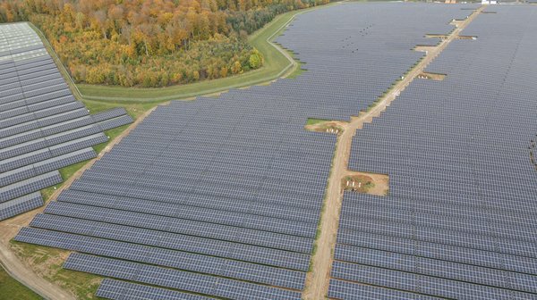 PV-Anlage Gickelfeld Luftaufnahme eines riesigen Solarparks mit unzähligen Solarpanelen, die sich über eine weite Fläche erstrecken. Ein sandiger Weg verläuft durch die Mitte der Anlage. Die Umgebung besteht aus grünen Wiesen und herbstlich gefärbten Wäldern mit gelben, orangen und roten Blättern.