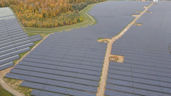 PV-Anlage Gickelfeld Luftaufnahme eines riesigen Solarparks mit unzähligen Solarpanelen, die sich über eine weite Fläche erstrecken. Ein sandiger Weg verläuft durch die Mitte der Anlage. Die Umgebung besteht aus grünen Wiesen und herbstlich gefärbten Wäldern mit gelben, orangen und roten Blättern.