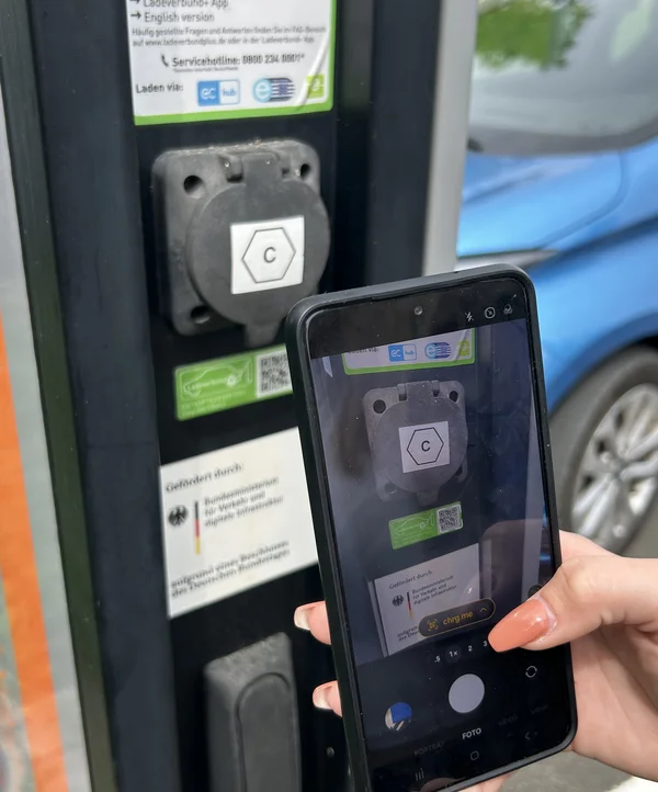 Hand hält ein Smartphone vor eine Ladestation, auf dem Bildschirm ist eine QR-Code-Scan-App geöffnet.