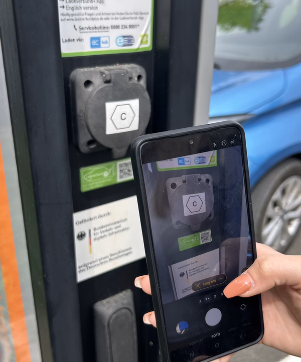 Hand hält ein Smartphone vor eine Ladestation, auf dem Bildschirm ist eine QR-Code-Scan-App geöffnet.