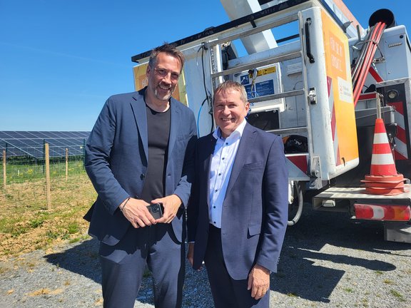 Zwei Männer im Anzug bei sonnigem Wetter vor einem Hubsteiger-Fahrzeug, im Hintergrund eine Photovoltaikanlage – symbolisch für den Einsatz erneuerbarer Energien.