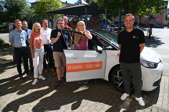 Eine Gruppe von acht Personen steht fröhlich neben einem weißen Elektroauto mit der Aufschrift „Carsharing – Schon dabei?“. Zwei Personen lehnen sich an die geöffnete Fahrertür, eine Frau hält einen Autoschlüssel in der Hand. Das Auto trägt die Logos von Stadtwerk Tauberfranken und einem Kooperationspartner. Im Hintergrund ist ein überdachter Bereich und eine Straße mit weiteren Passanten zu sehen. Die Szene spielt an einem sonnigen Tag auf einem gepflasterten Platz. Die Stimmung ist freundlich und gemeinschaftlich.