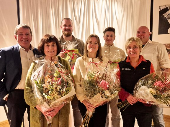 7 Personen stehen in schicker Kleidung vor einem weißen Vorhang. Vorne 3 Frauen mit einem Blumenstrauß in der Hand. Hinten 4 Männer. Die beiden in der Mitte halten einen großen Präsentekorb. Das Bild vermittelt die Situation einer Ehrung oder Beglückwünschung.
