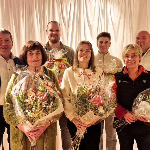Jubilare 2025 7 Personen stehen in schicker Kleidung vor einem weißen Vorhang. Vorne 3 Frauen mit einem Blumenstrauß in der Hand. Hinten 4 Männer. Die beiden in der Mitte halten einen großen Präsentekorb. Das Bild vermittelt die Situation einer Ehrung oder Beglückwünschung.