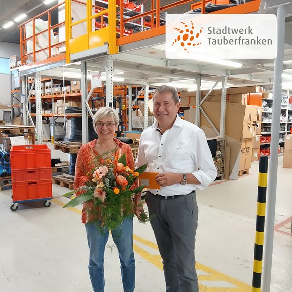 2 Personen stehen in einem Firmenlager mit Regalen. Beide sind leger gekleidet. Die blonde Frau links hält einen bunten Blumenstrauß in der Hand. Der Mann rechts im Bild übergibt die Blumen. Die Stimmung ist positiv.