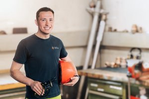 Mitarbeitender des Stadtwerks Tauberfranken steht in einer Werkstatt und hält einen orangefarbenen Schutzhelm sowie ein Messgerät in der Hand. Im Hintergrund sind Werkzeuge, Materialien und eine Werkbank unscharf zu sehen.