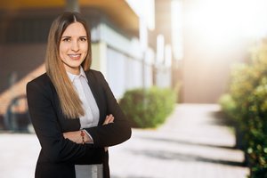 Mitarbeiterbild Junge Frau mit langen, glatten, dunkelblonden Haaren und einem Lächeln steht mit verschränkten Armen im Freien. Sie trägt eine weiße Bluse und einen schwarzen Blazer. Im Hintergrund ist ein sonniger, leicht unscharfer Außenbereich mit modernen Gebäuden und grünen Pflanzen zu sehen.