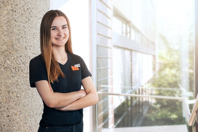 Eine junge Frau im schwarzen T-Shirt steht mit verschränkten Armen in einem Flur eines Geschäftsgebäudes. Der Flur ist lichtdurchflutet und die Frau lächelt in die Kamera. 