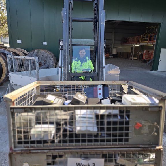Ein Mann in gelber Warnjacke sitzt auf einem Stabler in einem Werkshof. Im Hintergrund sind große Kabeltrommeln zu sehen sowie eine grüne Lagerhalle. Der Stapler hat vorne auf der Gabel zwei Gitterboxen mit alten Stromzählern.