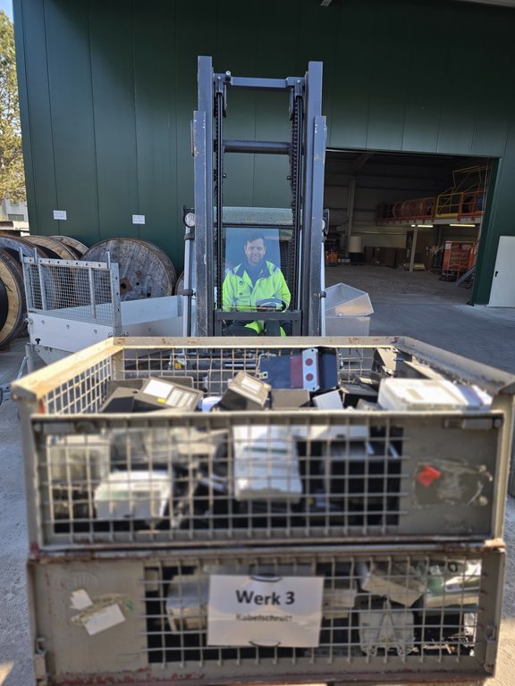 Ein Mann in gelber Warnjacke sitzt auf einem Stabler in einem Werkshof. Im Hintergrund sind große Kabeltrommeln zu sehen sowie eine grüne Lagerhalle. Der Stapler hat vorne auf der Gabel zwei Gitterboxen mit alten Stromzählern.
