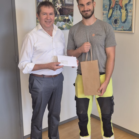 Zwei Personen stehen in einem Büro nebeneinander. Die Person links überreicht einen Umschlag, die Person rechts hält eine Geschenktasche. Im Hintergrund sind zwei gerahmte Bilder an der Wand zu sehen.