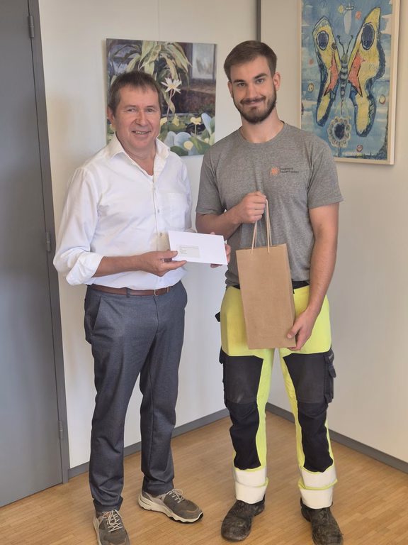 Zwei Personen stehen in einem Büro nebeneinander. Die Person links überreicht einen Umschlag, die Person rechts hält eine Geschenktasche. Im Hintergrund sind zwei gerahmte Bilder an der Wand zu sehen.