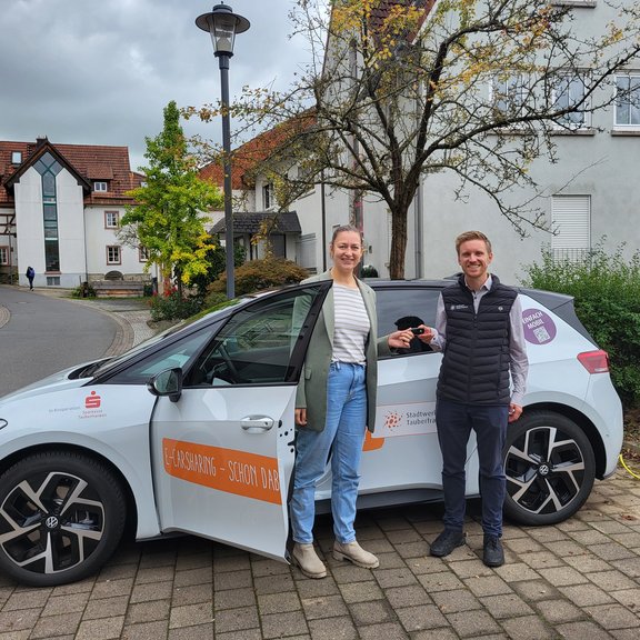 Ein weißes Elektroauto mit Werbeaufdruck vom Stadtwerk Tauberfranken in orange steht auf einem Platz in einer Innenstadt. Vor dem Auto stehen eine Frau und ein Mann. Ein Schlüssel wird übergeben. Die Sonne scheint. Der Platz ist leicht begrünt.