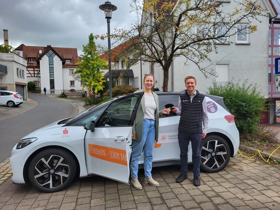 Ein weißes Elektroauto mit Werbeaufdruck vom Stadtwerk Tauberfranken in orange steht auf einem Platz in einer Innenstadt. Vor dem Auto stehen eine Frau und ein Mann. Ein Schlüssel wird übergeben. Die Sonne scheint. Der Platz ist leicht begrünt.