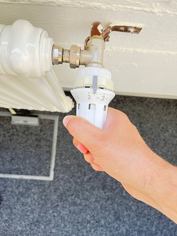 Eine Hand dreht an einem weißen Thermostat, das an einem Heizkörper befestigt ist.