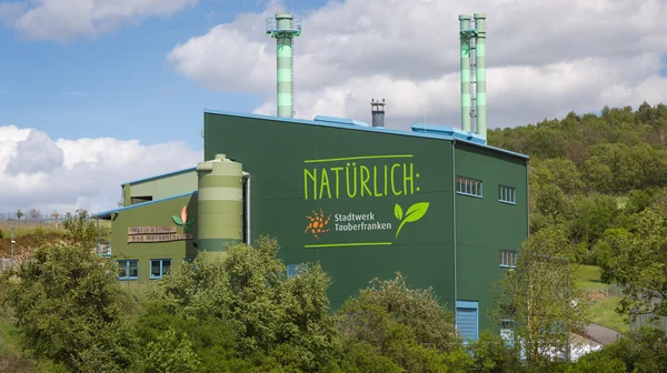 Grünes Industriegebäude mit der Aufschrift 'NATÜRLICH: Stadtwerk Tauberfranken' inmitten einer grünen Landschaft. Zwei grün-weiß gestreifte Schornsteine ragen in den Himmel. Auf der linken Seite ist ein weiteres Schild mit der Aufschrift 'Naturwärme Bad Mergentheim' zu sehen. Das Gebäude ist von Bäumen und Sträuchern umgeben, während der Himmel mit weißen Wolken bedeckt ist.