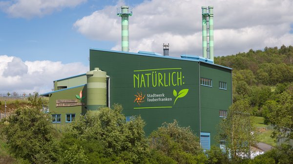 Naturwärmekraftwerk Grünes Industriegebäude mit der Aufschrift 'NATÜRLICH: Stadtwerk Tauberfranken' inmitten einer grünen Landschaft. Zwei grün-weiß gestreifte Schornsteine ragen in den Himmel. Auf der linken Seite ist ein weiteres Schild mit der Aufschrift 'Naturwärme Bad Mergentheim' zu sehen. Das Gebäude ist von Bäumen und Sträuchern umgeben, während der Himmel mit weißen Wolken bedeckt ist.