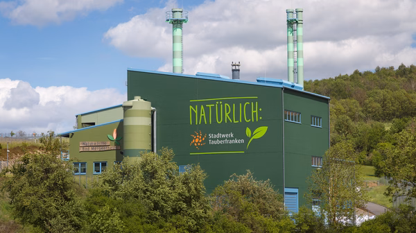 Naturwärmekraftwerk Grünes Industriegebäude mit der Aufschrift 'NATÜRLICH: Stadtwerk Tauberfranken' inmitten einer grünen Landschaft. Zwei grün-weiß gestreifte Schornsteine ragen in den Himmel. Auf der linken Seite ist ein weiteres Schild mit der Aufschrift 'Naturwärme Bad Mergentheim' zu sehen. Das Gebäude ist von Bäumen und Sträuchern umgeben, während der Himmel mit weißen Wolken bedeckt ist.