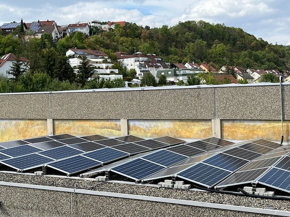 Solaranlage auf einem Flachdach mit mehreren Photovoltaik-Modulen, die Sonnenenergie nutzen. Im Hintergrund eine Wohnsiedlung mit Häusern auf einem grünen Hügel. Der Himmel ist teils bewölkt.