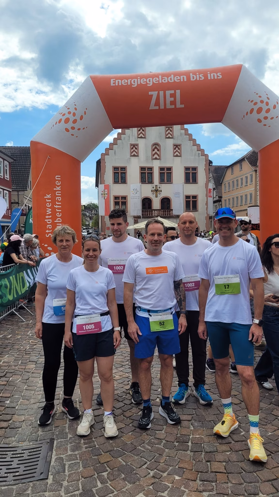 Stadtlauf Bad Mergentheim 2026 Mehrere Teilnehmende in weißen T‑Shirts und Laufschuhen stehen nach einem Lauf unter dem Zielbogen „Energieladen bis ins Ziel“ des Stadtwerks Tauberfranken, im Hintergrund ein historisches Rathaus auf einem Marktplatz.