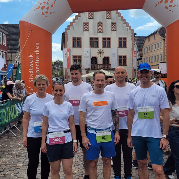 Mehrere Teilnehmende in weißen T‑Shirts und Laufschuhen stehen nach einem Lauf unter dem Zielbogen „Energieladen bis ins Ziel“ des Stadtwerks Tauberfranken, im Hintergrund ein historisches Rathaus auf einem Marktplatz.