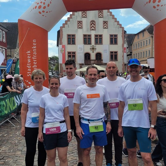 Mehrere Teilnehmende in weißen T‑Shirts und Laufschuhen stehen nach einem Lauf unter dem Zielbogen „Energieladen bis ins Ziel“ des Stadtwerks Tauberfranken, im Hintergrund ein historisches Rathaus auf einem Marktplatz.
