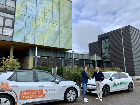 Zwei Männer stehen vor einem Gebäude mit moderner Architektur und schütteln sich die Hände. Links steht ein weißes Auto mit dem Aufdruck 'Aufladen statt Auftanken - Wir sind dabei. Stadtwerk Tauberfranken', rechts ein weiteres weißes Auto mit der Aufschrift 'SE Solar home'. Der Hintergrund zeigt ein farbenfrohes Wanddesign und bewölkten Himmel.