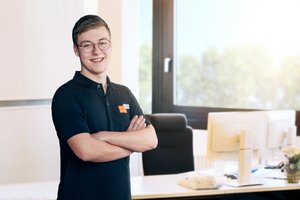 Mitarbeitender des Stadtwerks Tauberfranken steht in einem Büro mit verschränkten Armen vor einem Schreibtisch mit Monitoren. Im Hintergrund sind ein Fenster und unscharfe Arbeitsumgebung zu sehen.