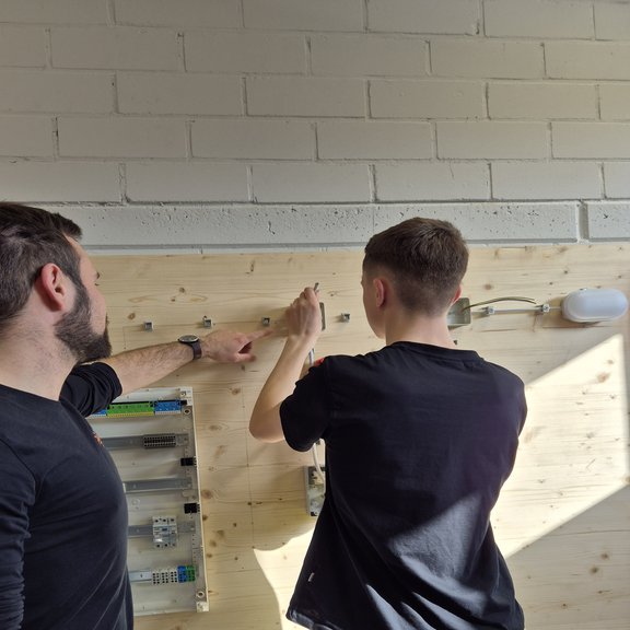 Ausbilder erklärt einem Auszubildenden den Aufbau einer elektrischen Schaltung an einer Übungswand in der Azubiwerkstatt.