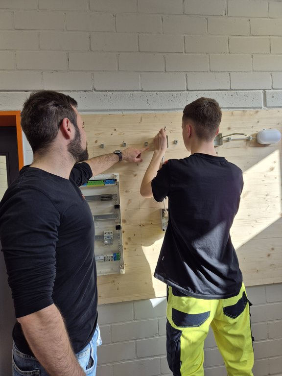 Azubiwerkstatt Ausbilder erklärt einem Auszubildenden den Aufbau einer elektrischen Schaltung an einer Übungswand in der Azubiwerkstatt.