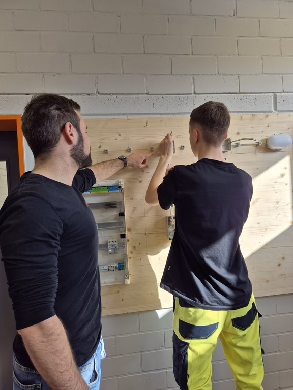 Azubiwerkstatt Ausbilder erklärt einem Auszubildenden den Aufbau einer elektrischen Schaltung an einer Übungswand in der Azubiwerkstatt.